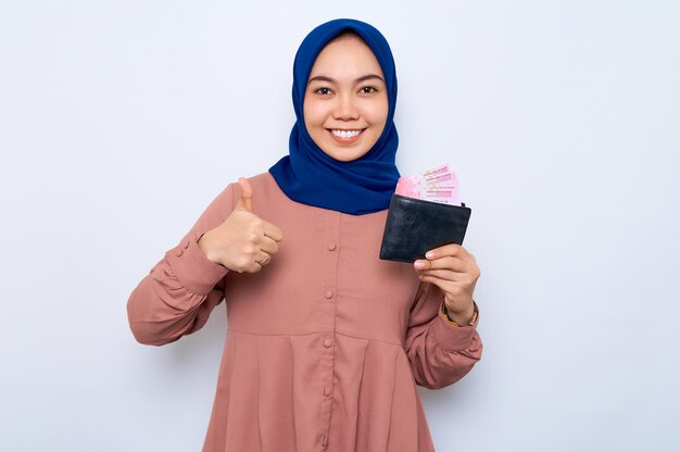 Smiling young Asian muslim woman in casual shirt holding wallet full of money banknotes and showing thumb up isolated over white background People lifestyle concept