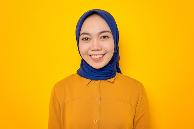 Smiling young asian muslim woman dressed in orange shirt\
looking at camera with happy face isolated on yellow\
background