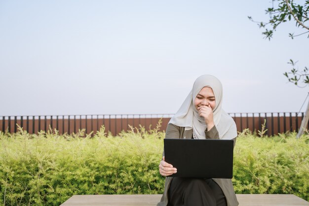 緑のスーツで若いアジアのイスラム教徒の女性を笑顔で公園でコンピューターに取り組んでいます。