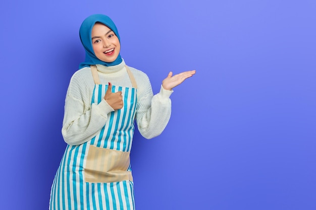 Smiling young Asian Muslim woman in hijab and striped apron pointing at copy space with  hands showing thumb up gesture isolated on purple background People housewife muslim lifestyle concept