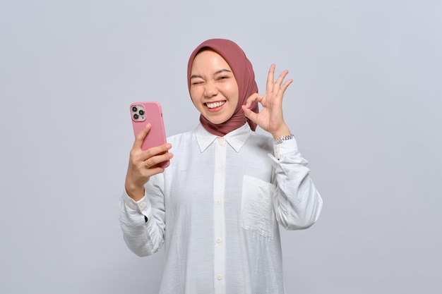 Улыбающаяся молодая азиатская мусульманка, держащая мобильный телефон и показывающая хороший знак на белом фоне