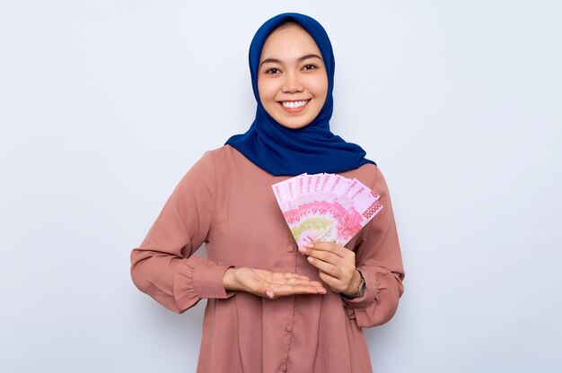 Улыбающаяся молодая азиатская мусульманка в розовой рубашке с денежными банкнотами, изолированными на белом фоне Концепция религиозного образа жизни людей
