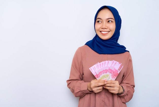 Smiling young Asian muslim woman in pink shirt holding money banknotes and looking aside isolated over white background People religious lifestyle concept