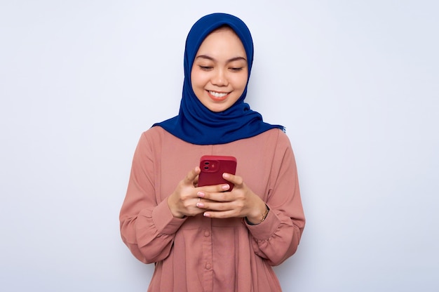 분홍색 셔츠를 입은 젊은 아시아 이슬람 여성이 휴대전화를 사용하여 웃고 있고 흰색 배경에 격리된 좋은 소식을 받았습니다.