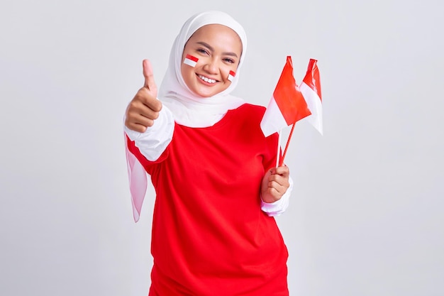 Smiling young Asian muslim woman in red white tshirt holding flag indonesian to celebrating indonesian independence day on 17 august and showing thumb up gesture isolated on white background