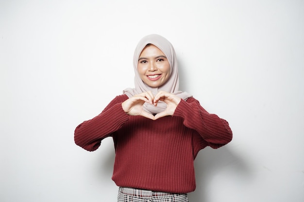 Улыбающаяся молодая азиатская мусульманка показывает сердечный жест, делая знак любви пальцами, изолированными на белом фоне