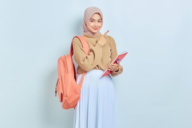 学校のコンセプトに戻って白い背景で隔離のコピースペースで脇に指を指しているバックパックと茶色のセーターで若いアジアのイスラム教徒の女性の学生を笑顔