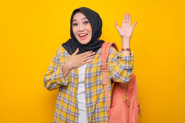 캐주얼 옷을 입고 배낭을 메고 웃고 있는 젊은 아시아 이슬람 여성 학생은 노란색 배경 교육 학교 대학 대학 개념에 고립된 누군가를 통지하면서 손으로 인사를 흔들었습니다.