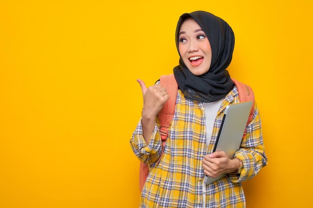 격자 무늬 셔츠와 배낭을 입은 웃고 있는 젊은 아시아 이슬람 여성 학생은 노란색 배경에 격리된 복사 공간에서 엄지손가락을 옆으로 가리키는 노트북 컴퓨터를 들고 있습니다.