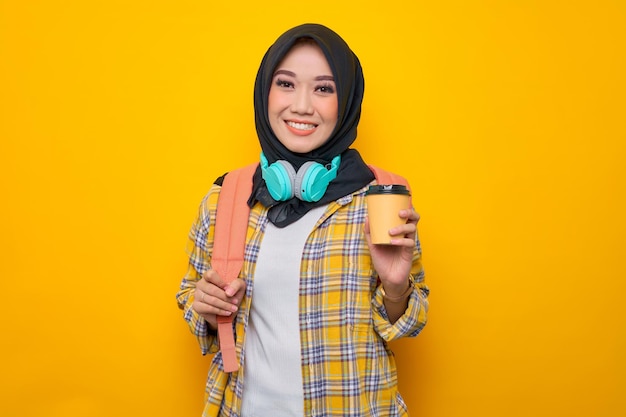 Smiling young Asian Muslim woman student in plaid shirt with headphones and backpack hold takeaway delivery cup coffee isolated on yellow background Education school university college concept