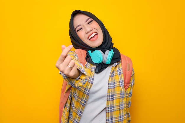 Smiling young asian muslim woman student in plaid shirt with\
headphones and backpack showing korean heart with two fingers\
crossed on yellow background education school university college\
concept