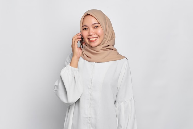 Smiling young Asian Muslim woman talking on mobile phone isolated over white background