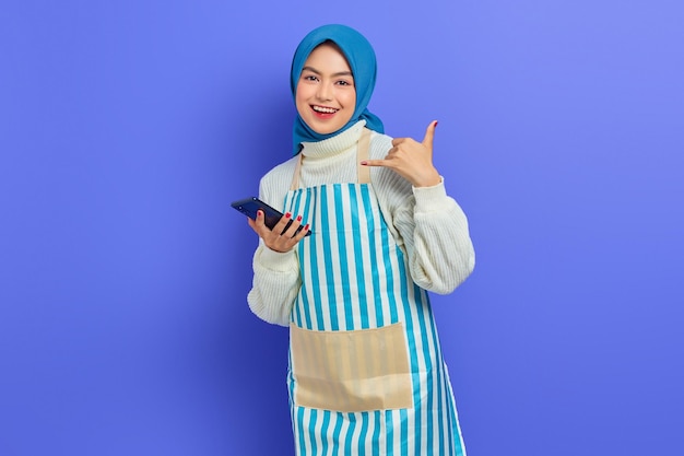 히잡과 앞치마를 입은 웃고 있는 젊은 아시아 이슬람 여성, 휴대전화를 들고 보라색 배경에 격리된 상태로 전화를 걸어달라는 전화 제스처를 하고 있습니다. 사람들이 주부 이슬람 라이프 스타일 개념