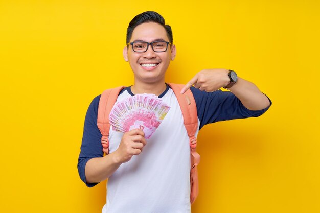 Smiling young asian student man in casual clothes and glasses backpack pointing finger at cash money isolated on yellow background education in high school university college concept