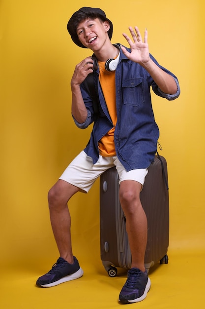 Foto sorridente giovane viaggiatore asiatico con abiti casual e cuffie che agita la mano verso la fotocamera dicendo