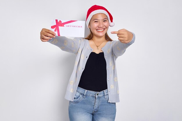 Smiling young Asian woman 20s in a Santa Claus hat receives and showing gift certificate coupon voucher card with finger isolated on white studio background Merry Christmas Concept