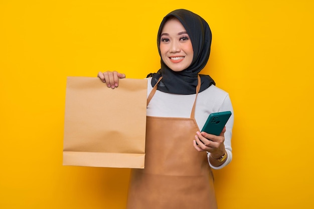 白いtシャツのエプロンで笑顔の若いアジアの女性バリスタは、携帯電話と白紙の持ち帰り用バッグを持ってコーヒーショップで働いて黄色の背景に分離されたモックアップビジネススタートアップ