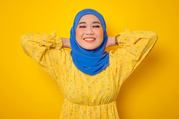 Smiling young asian woman in casual dress relaxing with hands\
behind head isolated on yellow background