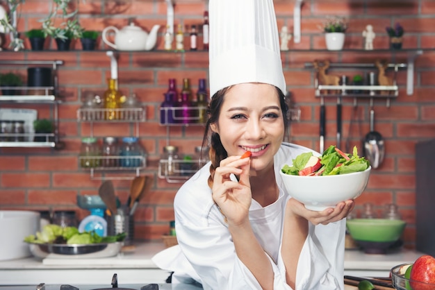 Smiling young Asian woman chef cook in white uniform standing at the kitchen, showing salad in bowl and red apple on her hand.