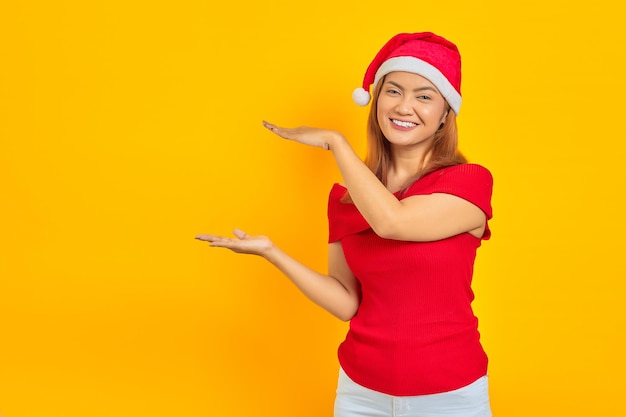 Smiling young Asian woman in christmas hat extending hand to the side inviting to come