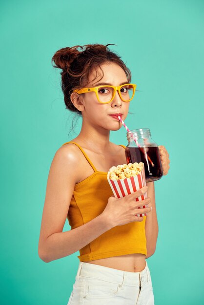 Smiling young Asian woman eating popcorn and drinking soda isolated over blue.