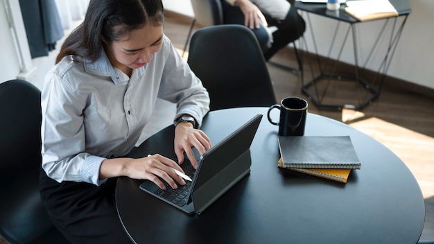 Foto sorridente giovane dipendente asiatica che si siede nella stanza dell'ufficio e utilizza il tablet del computer.