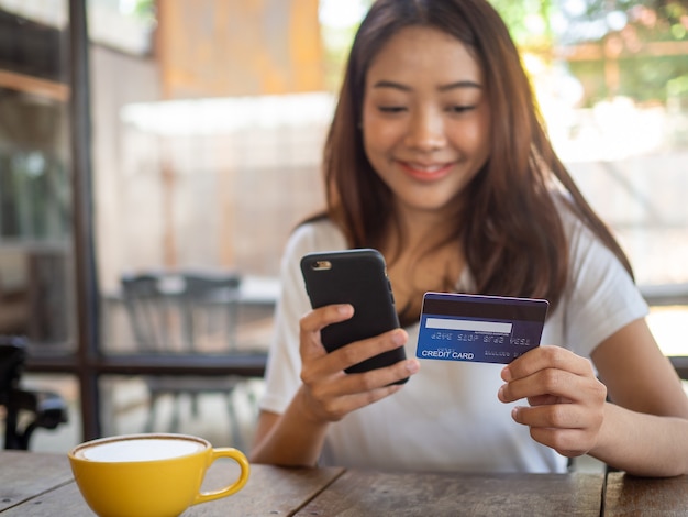 The smiling young Asian woman enjoys shopping online via a smartphone and paying online via credit card. Convenience of spending without cash. stay safe, shopping from home and Social distance