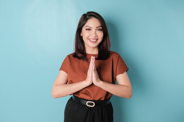 Smiling young Asian woman gesturing traditional greeting isolated over blue background