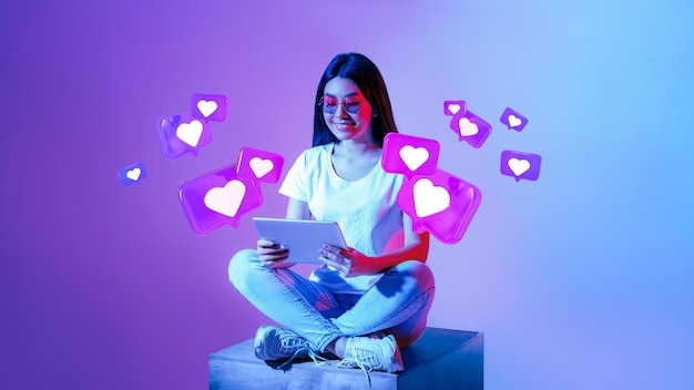 Smiling young asian woman in glasses typing on tablet has romantic chat with hearts on neon background