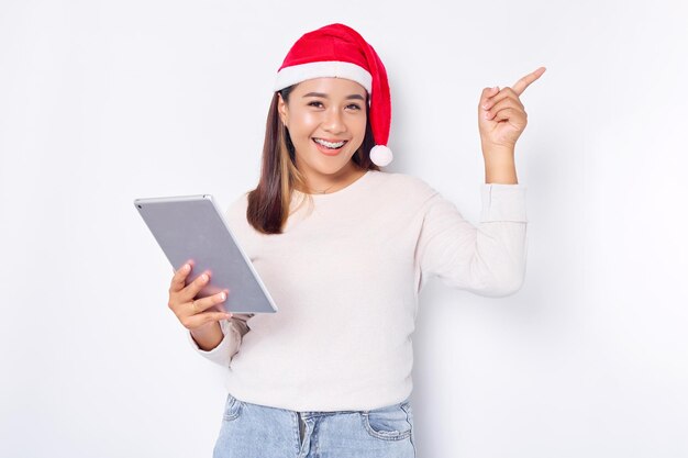 笑顔の若いアジア女性帽子クリスマス デジタル タブレットを保持し、白い背景の上に孤立した人差し指を脇にインドネシアの人々 を祝うクリスマス コンセプト