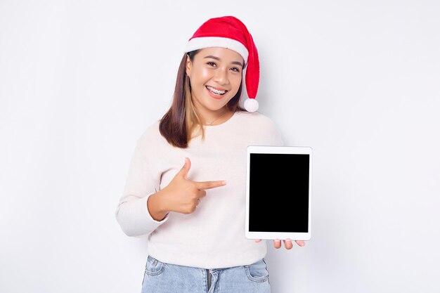 白い背景の上に孤立した空白の画面のタブレットで帽子クリスマス人差し指で笑顔の若いアジア女性インドネシアの人々 はクリスマスの概念を祝う