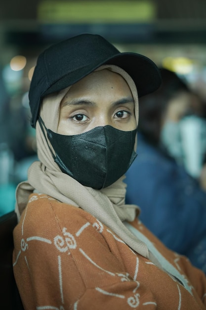 Smiling young asian woman in hijab wearing mask and hat sitting in waiting room