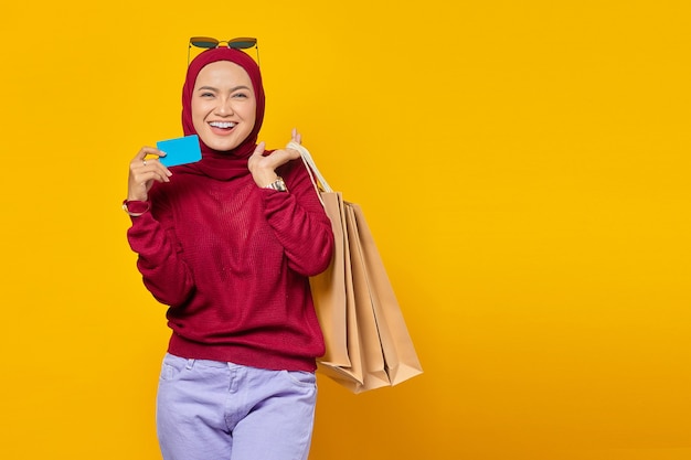 Smiling young asian woman holding credit card and shopping bags on yellow background