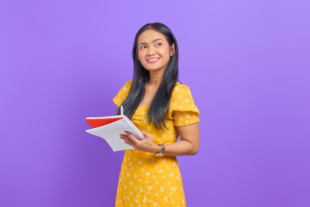 Smiling young Asian woman holding pen and notebook, looking up isolated on purple background
