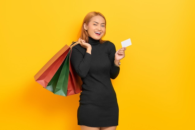 노란색 배경에 격리된 쇼핑백과 플라스틱 카드를 들고 웃고 있는 젊은 아시아 여성