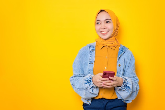 Smiling young Asian woman in jeans jacket holding mobile phone and looking aside at copy space isolated over yellow background