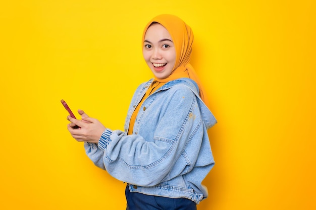 휴대 전화를 들고 노란색 배경에 고립 된 카메라를보고 청바지 재킷에 웃는 젊은 아시아 여성