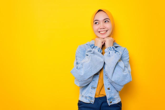 Smiling young Asian woman in jeans jacket looking up at copy space holding hands near chin isolated over yellow background