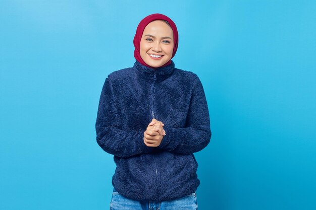 Smiling young Asian woman keeps hand together and feels optimistic on blue background