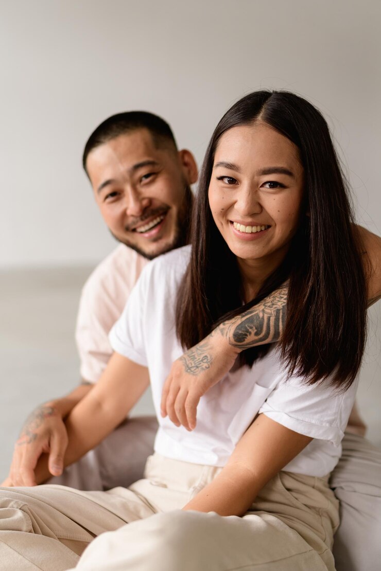 Photo 灰色の背景にタトゥーをした夫の近くに座ってカメラを見ている笑顔の若いアジア人女性