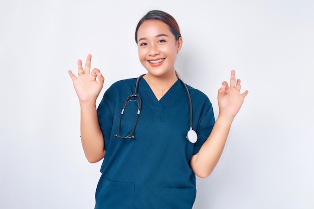 Smiling young Asian woman nurse wearing blue uniform with stethoscope assures the quality of test results in their clinic lab showing okay sign isolated on white background