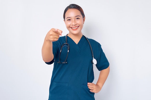 Smiling young Asian woman nurse wearing blue uniform with a stethoscope pointing finger at camera isolated on white background Healthcare medicine concept