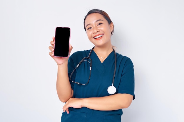 白い背景で隔離された空白の画面でスマートフォンを示す青い制服を着て働く笑顔の若いアジア女性看護師