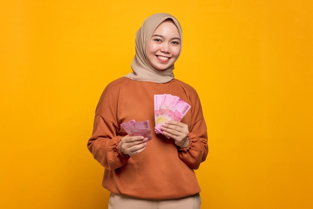 Smiling young asian woman in orange shirt giving money banknotes to someone isolated over yellow background