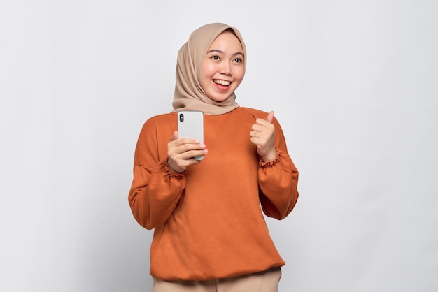 Smiling young Asian woman in orange shirt holding mobile phone and making thumb up gesture approve good thing isolated over white background