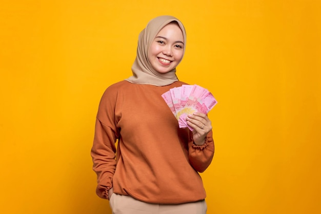 Smiling young asian woman in orange shirt holding money banknotes isolated over yellow background