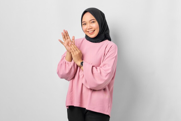 Smiling young Asian woman in pink shirt clapping in hands and looking at camera isolated over white background