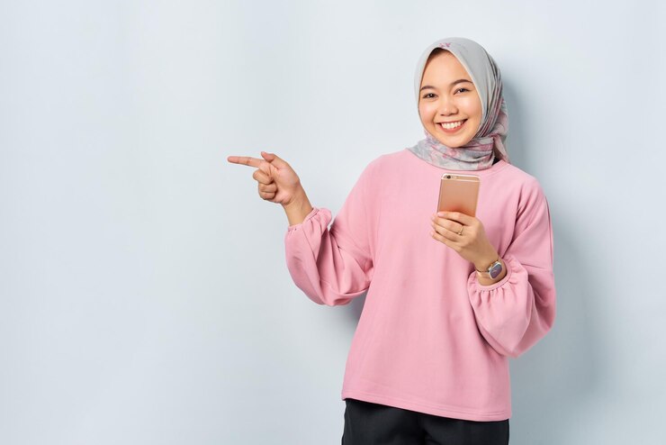 Smiling young Asian woman in pink shirt holding mobile phone and pointing finger away at blank space isolated over white background