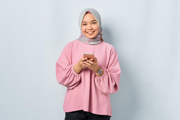 Smiling young Asian woman in pink shirt using mobile phone and looking at camera isolated over white background