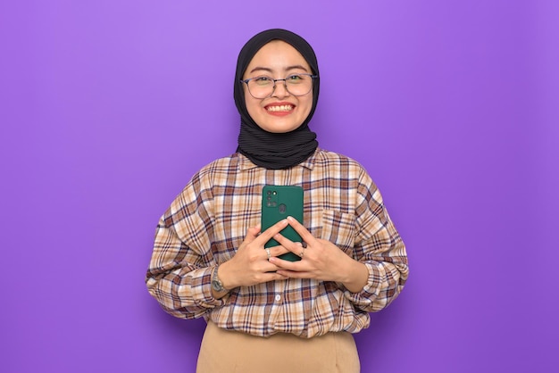 보라색 배경에 격리된 카메라를 바라보며 휴대폰을 들고 있는 격자 무늬 셔츠를 입은 웃고 있는 젊은 아시아 여성